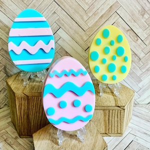 May include: Three pastel Easter eggs with different patterns. The eggs are on small stands and are arranged in a triangular shape. The egg on the left has blue and pink stripes and wavy lines. The egg in the middle has pink and blue wavy lines and dots. The egg on the right has yellow and green polka dots.