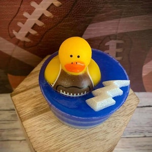 May include: A yellow rubber duck wearing a brown football jersey is sitting in a blue and white soap dish. The soap dish has a silver lightning bolt design.