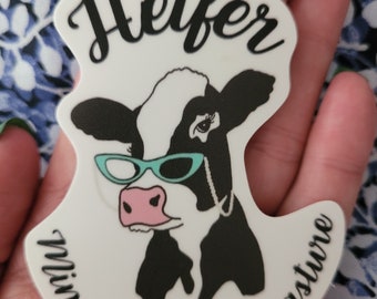 May include: White sticker with a black and white cow wearing blue sunglasses. The sticker says "Heifer Mind Your Own Pasture" in black text.