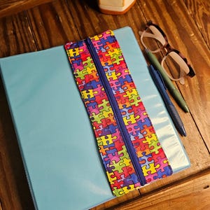 May include: A light blue binder with a colorful puzzle piece zipper pouch. The pouch has a blue zipper and is filled with multi-colored puzzle pieces. A pair of glasses and two pens are on the wooden surface.
