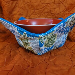 May include: A blue and white fabric bowl cozy with a floral pattern featuring seashells and starfish. The cozy is holding a red ceramic bowl.
