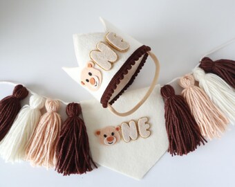 May include: A white felt crown with a brown felt trim and the word "ONE" in brown felt letters. A white felt bib with brown felt trim and the word "ONE" in brown felt letters. A brown, cream, and white yarn tassel garland.