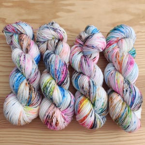 Könnte beinhalten: Vier Garnstränge in Weiß mit einem gesprenkelten, mehrfarbigen Muster mit leuchtenden Spritzern in Blau, Pink, Orange, Gelb und Schwarz. Jeder Strang ist zu einer traditionellen Form gedreht und zeigt ein einheitliches, buntes Farbmuster.