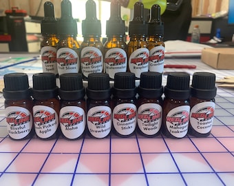 May include: A collection of 14 small brown glass bottles with black caps and white labels. Each bottle has a different scent, including Love Spell, Fruit Slices, Green Guava Pineapple, Watermelon, Raspberry, Cranberry, Blissful Blackberry, Fresh Picked Apple, Aloha, Wild Strawberry, Cinnamon Sticks, Twilight Woods, Mahogany Teakwood, and Toasted Coconut.