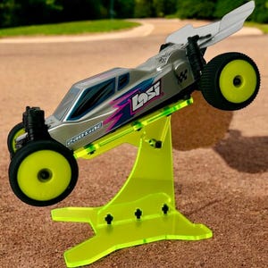 May include: A silver and black remote control car with a yellow and pink design is displayed on a bright yellow acrylic stand. The car is tilted forward and the stand has a unique shape.