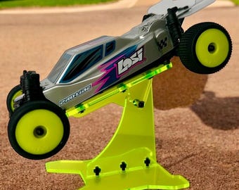 May include: A silver and black remote control car with a yellow and pink design is displayed on a bright yellow acrylic stand. The car is tilted forward and the stand has a unique shape.