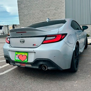 May include: A silver Toyota GR86 coupe with a black rear spoiler and a heart-shaped license plate frame. The car has a black rear bumper with dual exhaust tips.