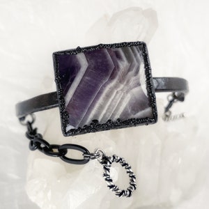 May include: A black metal bracelet with a square amethyst stone set in a black bezel. The bracelet has a chain detail and a small silver ring charm.
