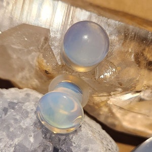 May include: Two iridescent blue glass ear gauges, one on top of the other, on a white crystal background.