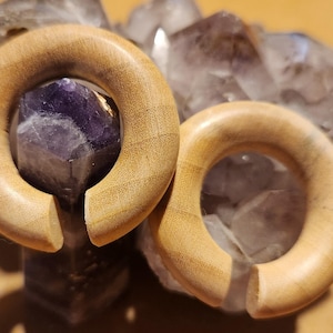 May include: Two wooden ear gauges, each with a circular shape and a gap on one side. The gauges are a light brown colour and are resting on a bed of purple and white crystals.