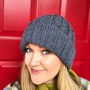 May include: A woman wearing a blue knitted beanie with a cable knit pattern. She is wearing a green turtleneck sweater and a blue and green patterned sweater over it.