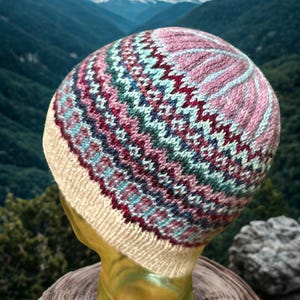 Peut inclure: Un bonnet tricoté avec un motif fair isle dans les tons de rose, sarcelle, bordeaux et crème. Le chapeau est présenté sur une tête de mannequin devant un fond de montagnes et un ciel nuageux. Le bord du chapeau est de couleur crème unie.