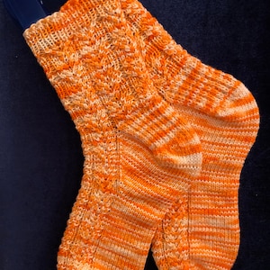 May include: A pair of hand-knitted socks in shades of orange and cream. The socks feature a textured pattern on the leg and a striped pattern on the foot. The socks are displayed on a blue hanger against a dark blue background.