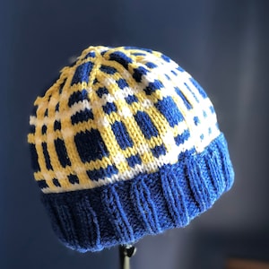 May include: A knitted beanie hat with a blue brim and a pattern of yellow and white squares. The hat is made of a soft, textured yarn and is designed to keep the wearer warm. The hat is a stylish accessory for cold weather.