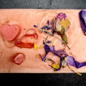 May include: A pink soap bar with dried flowers and heart-shaped embellishments. The soap has a textured surface and is decorated with dried lavender, rose petals, and other botanicals.