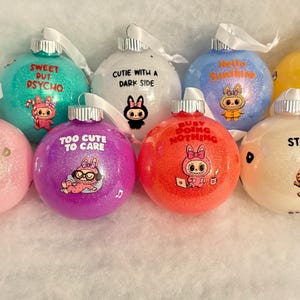 以下が含まれることがあります： さまざまな色のキラキラ光るクリスマスオーナメントのコレクション。それぞれに漫画のキャラクターと「Stay Wild」や「Too Cute to Care」などのユニークなフレーズが描かれています。オーナメントにはシルバーのキャップと白いリボンが付いています。