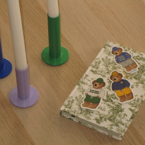 May include: Three candle holders with different colored bases: blue, purple, and green. The candle holders are on a wooden surface. A notebook with a white cover and green floral pattern is in the background. The notebook has three bear stickers on it.
