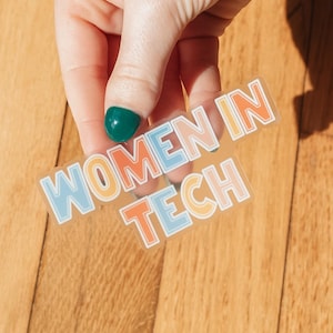 May include: A clear sticker with the text "WOMEN IN TECH" in a colorful, retro font. The sticker has a white outline and is held against a wooden surface. The letters are in shades of blue, orange, and peach.