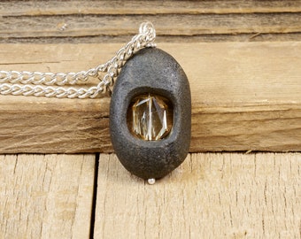 May include: A silver chain necklace with a pendant made from a dark gray stone with a cut-out window revealing a clear, faceted, amber-colored crystal.