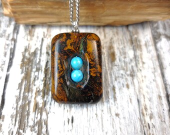 May include: A brown and gold rectangular pendant with two turquoise stones set in the center. The pendant is hanging from a silver chain.