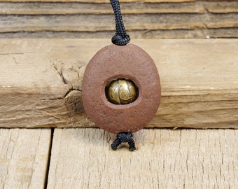 May include: A brown clay pendant with a hole in the center, revealing a gold-colored metal bead. The pendant is attached to a black cord.
