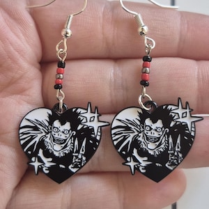 May include: A pair of heart-shaped earrings featuring a black and white anime character design. The earrings have silver-colored hooks and are accented with black and red beads. The character is holding two knives.