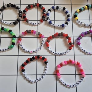 Op de afbeelding: Een verzameling kralenarmbanden in verschillende kleuren, waaronder zwart, rood, groen en paars. Elke armband heeft witte letterkralen met woorden als "ASHES", "RATS" en "SEE THE LIGHT". De armbanden zijn gerangschikt op een wit raster.