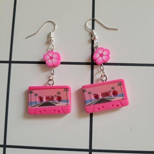 Peut inclure: Une paire de boucles d'oreilles en forme de cassette rose avec un charme de fleur rose. Les cassettes ont un motif de palmier et un fond rose.