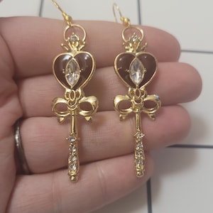 Op de afbeelding: Een paar gouden oorbellen met een hartvormige hanger, een kroon, een strik en een sleutelontwerp. De hartvormige hanger heeft een bruine emaille afwerking en een heldere strass in het midden. Het sleutelontwerp is versierd met heldere strass.