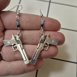 May include: Silver-toned gun-shaped earrings with intricate detailing. Each earring features a small chain with blue and gold beads. The earrings are designed with a hook closure for pierced ears.