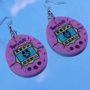 Może przedstawiać: Para r&oacute;żowych kolczyk&oacute;w Tamagotchi. Każdy kolczyk ma r&oacute;żowy owalny kształt z ż&oacute;łtym ekranem wyświetlającym pikselową postać. Kolczyki mają wz&oacute;r posypki na krawędziach, a słowo "Tamagotchi" jest wydrukowane na g&oacute;rze.