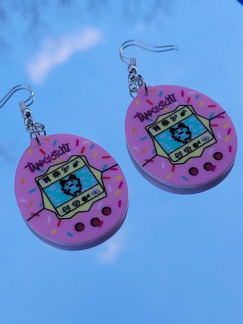 Może przedstawiać: Para r&oacute;żowych kolczyk&oacute;w Tamagotchi. Każdy kolczyk ma r&oacute;żowy owalny kształt z ż&oacute;łtym ekranem wyświetlającym pikselową postać. Kolczyki mają wz&oacute;r posypki na krawędziach, a słowo "Tamagotchi" jest wydrukowane na g&oacute;rze.