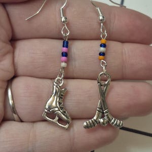 May include: Silver-toned hockey-themed earrings. One earring features a skate charm, the other crossed hockey sticks. Each earring has a beaded accent with blue, pink, and white beads, and orange, blue, and yellow beads.