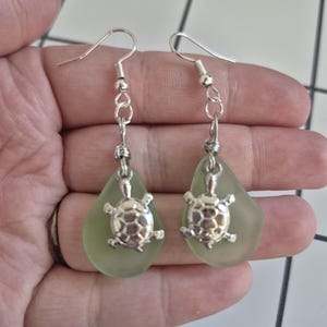 Puede incluir: Un par de pendientes con un dije de tortuga plateada suspendido de una cuenta de vidrio en forma de lágrima verde claro. Los pendientes tienen ganchos y cadenas plateadas. Fondo blanco y gris.