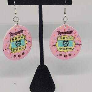 Może przedstawiać: R&oacute;żowe kolczyki Tamagotchi ze srebrnymi haczykami. Kolczyki mają r&oacute;żowe tło z kolorowymi posypkami i napisem "Tamagotchi". Środek kolczyk&oacute;w ma ż&oacute;łty ekran z pikselową postacią.