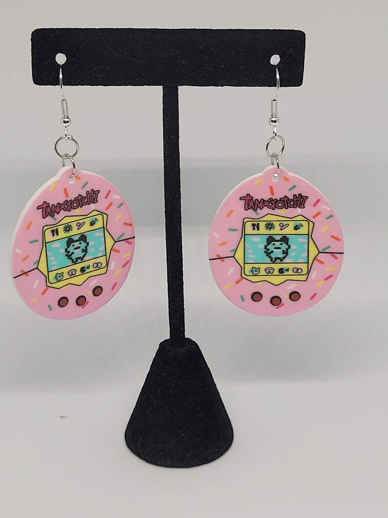Może przedstawiać: R&oacute;żowe kolczyki Tamagotchi ze srebrnymi haczykami. Kolczyki mają r&oacute;żowe tło z kolorowymi posypkami i napisem "Tamagotchi". Środek kolczyk&oacute;w ma ż&oacute;łty ekran z pikselową postacią.