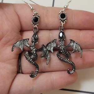 May include: A pair of silver dragon earrings with black crystal accents. The dragons are detailed with wings and curled tails.