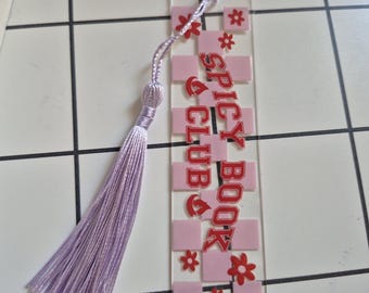 Peut inclure: Marque-page transparent en acrylique avec un motif à carreaux roses et blancs. Le texte "SPICY BOOK CLUB" est en rouge. Il est orné d'un pompon lavande attaché en haut avec un cordon assorti et une petite boucle.