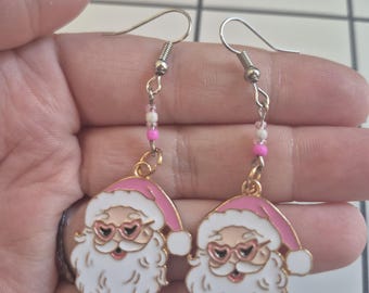 May include: Dangle earrings with Santa Claus faces, pink hats, and heart-shaped glasses. The earrings have white beards and are accented with pink and white beads. The hooks are silver-coloured.