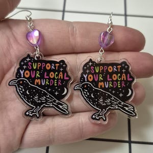 May include: A pair of dangle earrings with a black crow design. The earrings have a heart-shaped bead in purple and the text "SUPPORT YOUR LOCAL MURDER" in colorful letters.