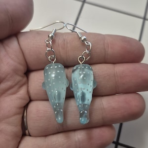 May include: A pair of light blue, translucent earrings shaped like small whales. Each earring features white polka dots and silver-colored fish hook ear wires. The earrings are held in a hand.