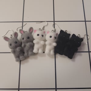 Peut inclure: Quatre paires de boucles d'oreilles pendantes avec de petites figurines d'animaux en peluche. Deux paires sont des lapins gris, une paire est des chatons blancs et une paire est des chatons noirs.