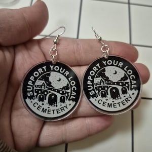 May include: A pair of round black and white earrings with the text "Support Your Local Cemetery". The earrings feature a cemetery scene with a full moon, a house, and tombstones. The earrings have silver-colored hooks.