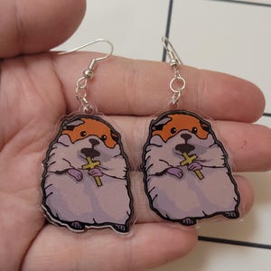 May include: A pair of clear acrylic earrings featuring a cartoon hamster wearing a purple robe and holding a gold cross.