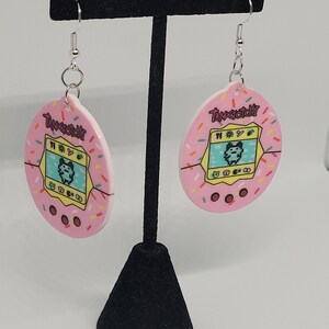Może przedstawiać: R&oacute;żowe kolczyki Tamagotchi z posypką. Każdy kolczyk ma retro cyfrowy wz&oacute;r zwierzaka z napisem "Tamagotchi". Kolczyki wiszą na srebrnych haczykach.