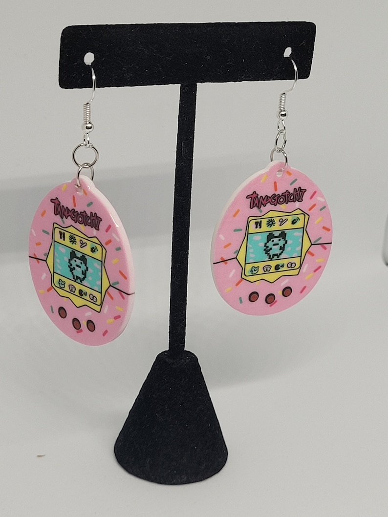 Może przedstawiać: R&oacute;żowe kolczyki Tamagotchi z posypką. Każdy kolczyk ma retro cyfrowy wz&oacute;r zwierzaka z napisem "Tamagotchi". Kolczyki wiszą na srebrnych haczykach.