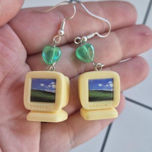 May include: A pair of dangle earrings featuring miniature beige computer monitors with a screen graphic of a green hill and blue sky. Each earring is accented with a small, translucent green heart-shaped bead and a silver-toned metal hook.