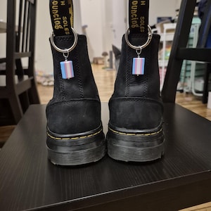 Pode incluir: Um par de botas pretas com solas grossas. Cada bota tem uma presilha de tecido preto com as palavras "WITH BOUNCING SOLES" em amarelo. Um pequeno pingente com as cores da bandeira transgênero pende de um anel prateado em cada bota.