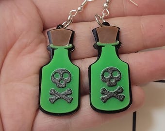 Peut inclure: Une paire de boucles d'oreilles en forme de bouteille de poison verte avec un contour noir. Chaque boucle d'oreille présente un motif de tête de mort et d'os croisés noirs. Les boucles d'oreilles ont des crochets d'oreille argentés et un bouchon marron.