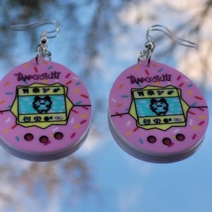 Może przedstawiać: Para r&oacute;żowych kolczyk&oacute;w Tamagotchi. Każdy kolczyk ma r&oacute;żowy owalny kształt z ż&oacute;łtą obw&oacute;dką i kolorową posypką. Ekran Tamagotchi wyświetla spikselizowanego zwierzaka. Srebrne haczyki do uszu dopełniają design.